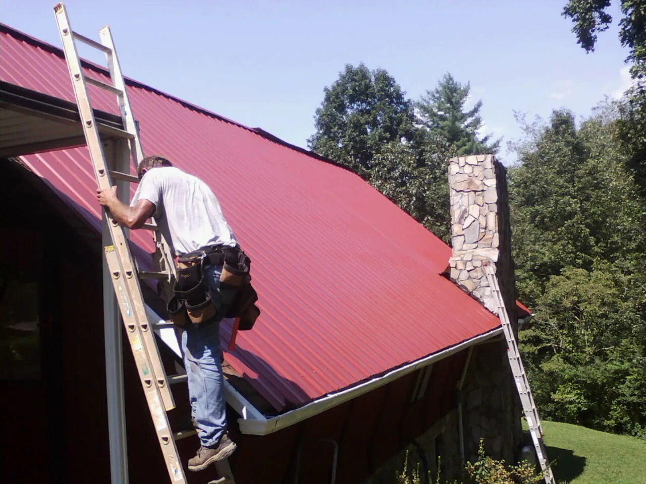 Roofer on a ladder beside a red metal roof and stone chimney — Soffit & Fascia Repair by Roofing Brothers in Grapeville